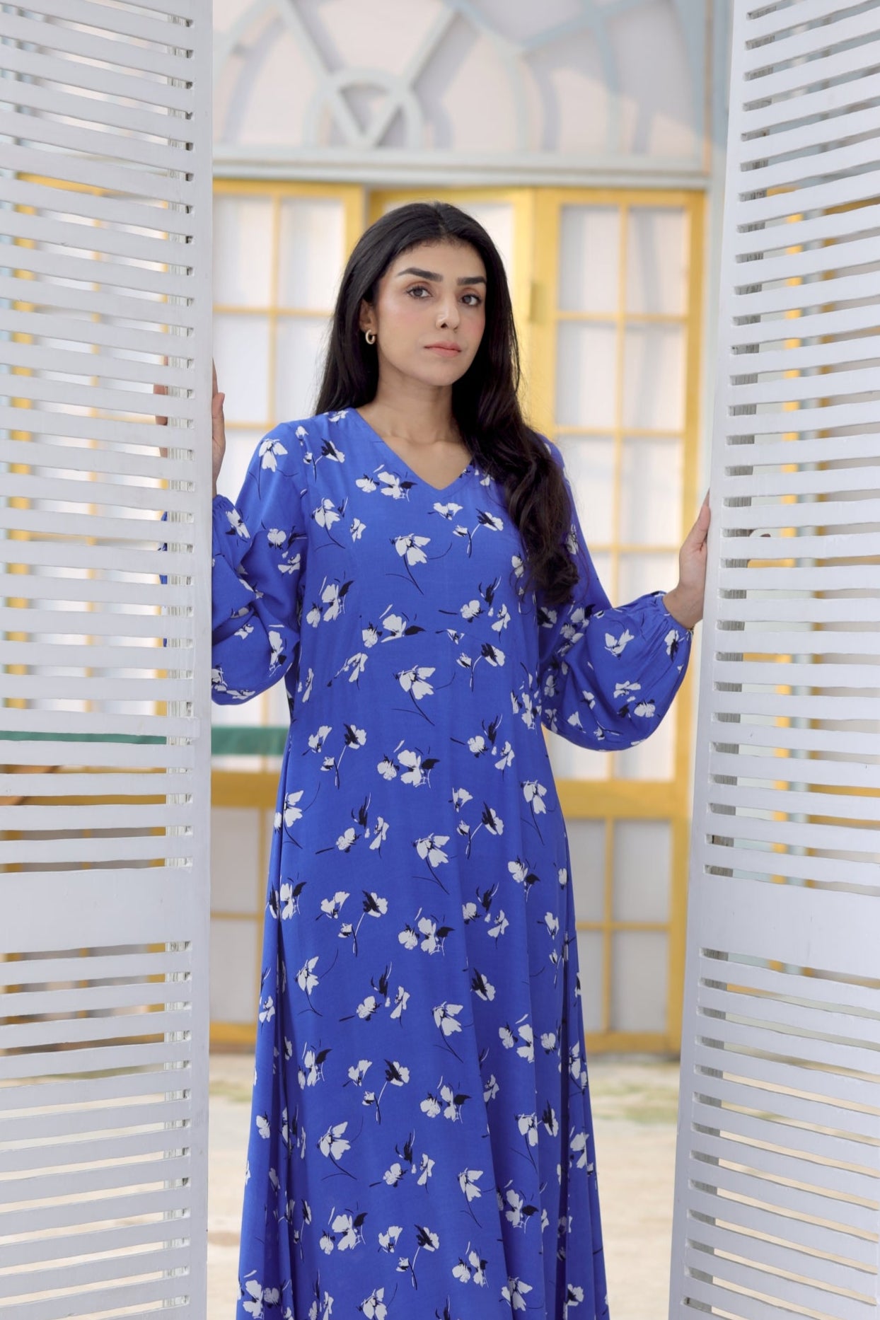 Woman wearing a blue dress with white floral pattern standing in front of a white lattice door.